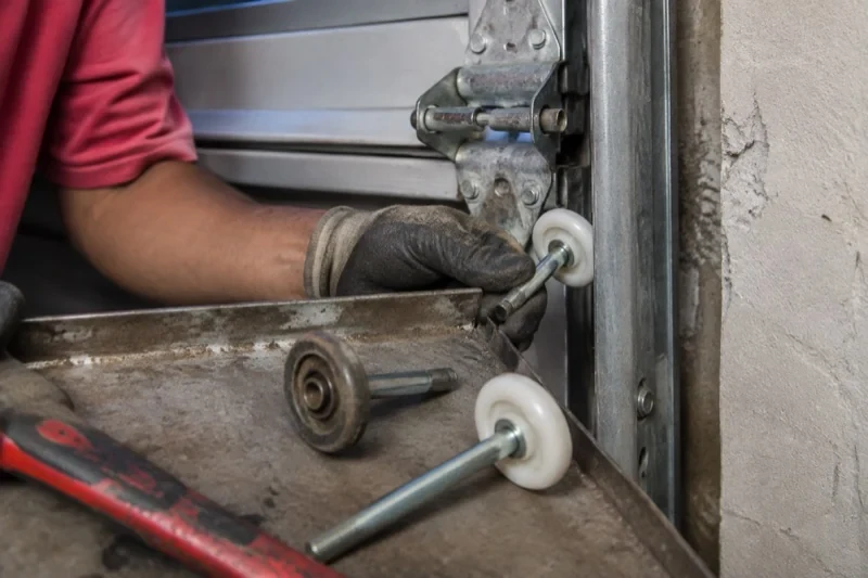 Gloved hand holding a white garage-door roller beside its track, with two loose rollers and a red tool on a tray.